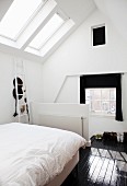 Bed with white bed linen in front of half-height balustrade wall in attic room with skylight and dark wooden floor