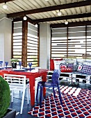 Roofed terrace with old table, collection of chairs and rug with red, white and blue pattern in front of armchairs upcycled from old pallets with scatter cushions