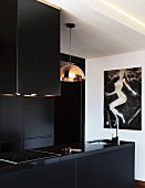 Black kitchen counter with sink and hob below extractor hood next to pendant lamp with copper-coloured lampshade