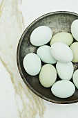 A pewter bowl filled with blue eggs on a white marble surface