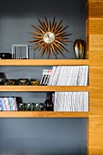 Collection of Murano glass ornaments and books on wooden shelves against grey wall below sunburst-shaped wall clock