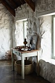 Vintage Holztisch an weiss getünchter, rustikaler Steinwand in hellem Raum; unter dem Tisch ein großer Korb
