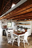 Offener Wohnraum mit einfacher Küche und Holztisch mit weissen Stühlen unter rustikaler Holzbalkendecke