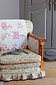 Traditional armchair with floral upholstery and embroidered antimacassar on backrest in front of farmhouse cupboard painted grey