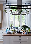 Crockery and food on island counter below stainless steel extractor hood with posies and lit candle lanterns hanging from suspended metal rack