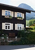 Dunkles Holzhaus mit Blumenkästen an Fenster und helle Holzläden, im Hintergrund Berglandschaft