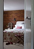 View through open door into bedroom with floral bedspread on bed against rustic wooden wall