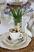 Österliches Gedeck auf Kaffeetafel mit Vogelnest und Keks-Hase in der Tasse und weißem Porzellanhase; Traubenhyazinthen in Marmeladenglas im Hintergrund