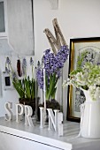 Shabby chic arrangement on white cabinet; spring flowers in glass vessels and enamel can, gilt-framed engraving and letters reading SPRING