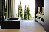 Black bathtub opposite fitted washstand in front of glass wall with view of group of trees