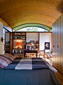 Bedroom with checked bedspread on double bed below wood-clad barrel-vaulted ceiling: antique wooden table and bookcases in background