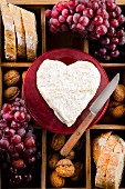 A heart-shaped Neufchatel cheese on a wooden crate with bread, nuts and red grapes