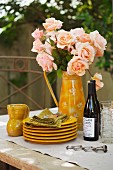 Stacked plates, salmon pink roses and bottles of wine on table in garden