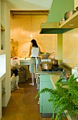 Mattgrüner Vintage Gasherd mit Kupfergeschirr in Landhausküche; Hund und Frau beim Gemüseputzen an langem Spülbecken