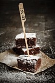 Three brownies on brown paper dusted with icing sugar and an Eat Me sign