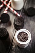 An Oreo biscuit in a glass of milk, drinking straws and Oreo biscuits on a wooden slab