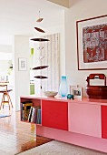 Sideboard painted pink and red with books in open cupboard below wooden mobile