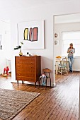 Modern artwork above chest of drawers against wall between two doorways in interior with continuous wooden floor; woman in front of window in background