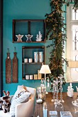 Glass candle sticks, table lamp and candle lanterns on table in front of Christmas decorations in wooden display case and garland of fir branches and fairy lights on turquoise wall
