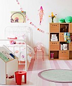 Simple writing desk, red stool, white metal bed, small plastic chair, wooden storage boxes on shelves and round rug on striped carpet