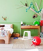 Modern wooden bed next to writing desk and white plastic stool below stylised tree made from wooden planks on green wall; red space hopper on rug in foreground