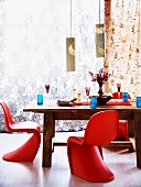 Rote Panton Stühle um massiven Holztisch mit blauen Trinkgläsern gedeckt und blumige zarte Vorhänge im Hintergrund