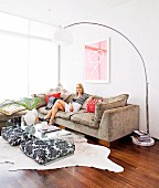 Arc lamp, woman sitting on sofa and black and white patterned floor cushions under transparent coffee table in comfortable, sunny lounge area