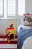 Detail of child's bedroom - toys on yellow-painted Thonet chair below window with closed white interior shutters and bed with patterned bedspread