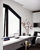 White, continuous desk and Eames chair below window with dark, sloping frame