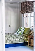 Blick durch offene Tür auf Bett mit gemusterter Tagesdecke und Kissenstapel, dahinter eingebauter Schrank in weißem, holzverkleidetem Zimmer
