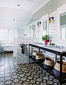 Spacious bathroom with black and white patterned floor tiles, modern washstand with marble top on black metal frame next to shower area and free-standing bathtub below window