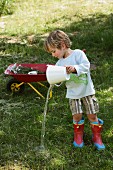 Kind in Gummistiefeln & mit selbstgenähtem Shirt spielt mit Wassereimer im Garten