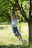 Junge mit selbstgenähtem Tiermotiv auf Shirt schaukelt an Ast eines Baumes im Garten
