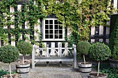 Weathered wooden bench flanked by rows of potted box trees in front of climber-covered half-timbered house with lattice windows