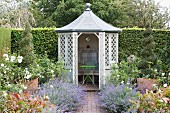 Path lined with flowers leading to white, wooden latticed summer house with pavilion roof with open door and view of green-painted table