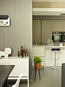 Detail of dining area with white sideboard against wooden wall varnished pale grey; modern, white fitted kitchen with bar stools at counter in background