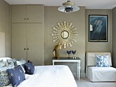 Bedroom in shades of grey & blue with bedroom, floor-to-ceiling wardrobe, mirror with sunburst frame, side table and easy chair in niche