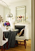 Dining area with grey damask tablecloth on round table and designer arc lamp; vintage mirror above open fireplace in background