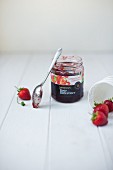 A jar of supermarket strawberry jam with a spoon and fresh strawberries.