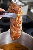 a hand holds a tenderloin beef above a pot with pieces of carrots and onions frying, for a typical Czech dish called Svickova
