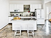 Bar stools at central island with stainless steel worksurface in white, open-plan kitchen