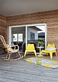 Wicker rocking chair, yellow plastic chairs and long cushion on floor of roofed wooden terrace