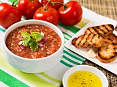 Gazpacho, garniert mit Avocado, roten Zwiebeln und Basilikum, daneben Olivenöl, Crostini und frische Tomaten