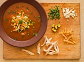 Tortilla soup on a chopping board with garnishes (chicken, tortilla strips, avocado, coriander and cheese)