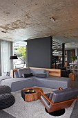 Gemütliche elegante Designer-Sofalandschaft mit grauen Polstern vor dunkelgrauer Wandscheibe in offenem Wohnbereich mit Betondecke