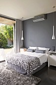 Elegantes graues Polster-Doppelbett vor dunkelgrauer Wand und zwei weißen Designer-Pendelleuchten mit Blick auf begrünte Terrasse