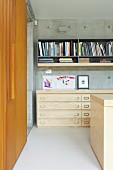Half-height pale wooden chest of drawers below grey shelves on concrete wall and wooden sliding doors to one side in modern interior