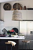 Wicker lamps above marble kitchen counter, books stacked next to fruit bowl and African basket on white wall units in background