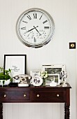 Several black and white photos in silver frames and vintage telephone on elegant console table below clock with Roman numerals on wall