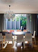 Kugelförmige Hängeleuchte Dandelion von Richard Hutten über Esstisch mit Designer-Stühlen aus hellem Kunststoff; Blick auf Terrasse mit blaugrauer Sichtschutzwand